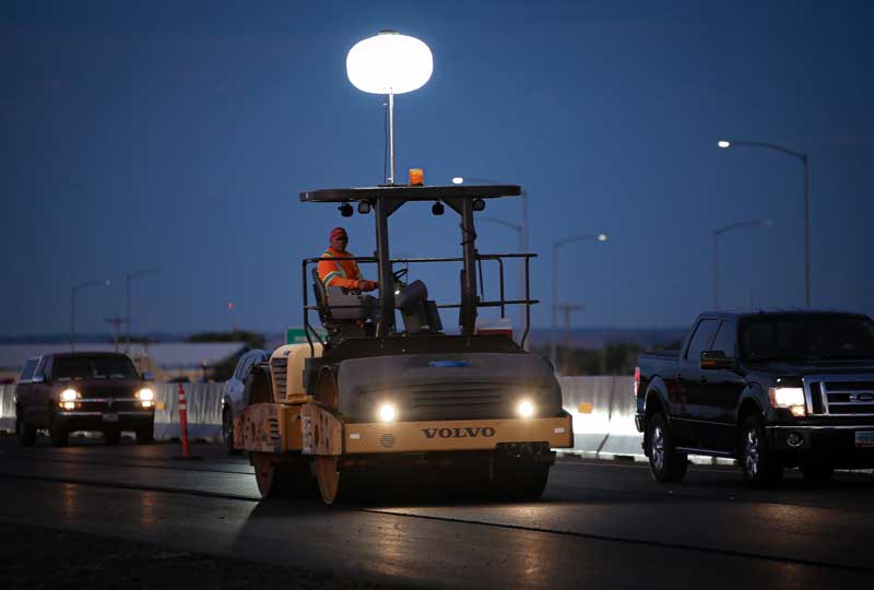 A worker operates a roller to smooth the new asphalt surface in the closed lane.
